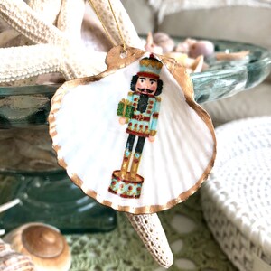 May include: A seashell Christmas ornament featuring a painted nutcracker soldier. The white shell has a gold-painted edge and a gold string for hanging. The nutcracker is in a blue and gold uniform, holding a green gift.