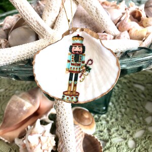 May include: A hand-painted Christmas ornament featuring a nutcracker soldier on a scallop shell. The shell has a gold-painted edge and a gold string for hanging. The nutcracker is in a blue uniform and holds a candy cane.