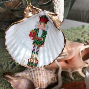 May include: A Christmas ornament made from a white scallop shell, featuring a painted nutcracker soldier holding a gift. The shell's edge is trimmed with gold paint, and a gold string is attached for hanging. The background includes seashells and greenery.