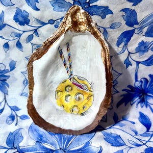 May include: An oyster shell painted with a gold rim and a white interior. The shell features a yellow circle with a dotted pattern and a red and purple design, hanging from two colorful strings. The shell is on a blue and white floral patterned surface.