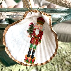 May include: A Christmas ornament made from a white seashell, featuring a painted nutcracker soldier holding a Christmas tree. The shell has a gold-painted edge and a gold string for hanging. The nutcracker is dressed in a red and green uniform.