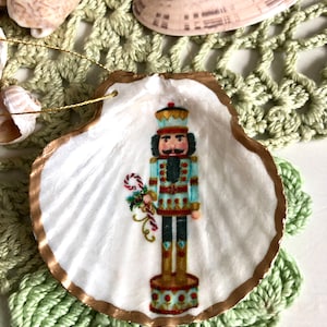 May include: A seashell ornament featuring a painted nutcracker soldier. The shell has a gold-painted edge and a white interior. The nutcracker is in a blue and red uniform, holding a candy cane and flowers. The ornament is on a green crocheted surface.