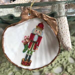 May include: A seashell Christmas ornament featuring a painted nutcracker soldier. The shell is white with a gold-painted edge and a gold string for hanging. The nutcracker is dressed in a red coat and black boots, holding a stocking.
