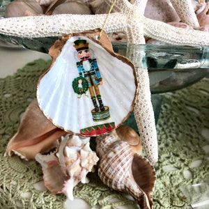 May include: A seashell Christmas ornament featuring a painted nutcracker soldier in blue, gold, and green. The shell has a gold-painted edge and a gold string for hanging. Other seashells and a starfish are in the background.