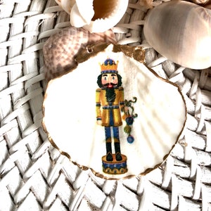May include: A white seashell decorated with a gold-edged image of a nutcracker soldier in a yellow and blue uniform, holding a cluster of ornaments. The shell is surrounded by other seashells on a white woven surface.