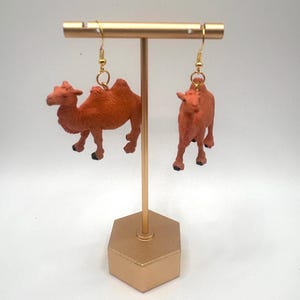 Può includere: Un paio di orecchini pendenti con miniature di cammelli color arancione con zoccoli neri. I cammelli sono appesi a ganci in metallo dorato. Gli orecchini sono esposti su un supporto a forma di T color oro con una base esagonale.