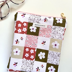 May include: A patchwork zippered pouch with a floral pattern in white, pink, and brown squares. The pouch has a gold zipper pull and the text "blueberry & blooms". Eyeglasses are in the upper left corner.