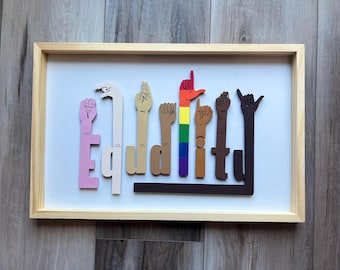 May include: Wooden framed artwork with the word "Equality" spelled out in colorful hand gestures. The hands are various colors, including pink, tan, brown, and rainbow, against a white background. The frame is a light wood color.