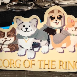 May include: A sticker featuring four cartoon corgi dogs dressed in fantasy attire. The sticker has the text "CORG OF THE RINGS" in gold lettering. The corgis are in various colors, including brown, white, and orange.