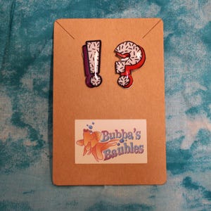 May include: Two enamel pins on a brown card. One pin is an exclamation point, the other a question mark, both with a white and black speckled design. The card has a sticker with a goldfish and the text "Bubba's Baubles".