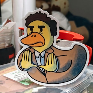 Puede incluir: Una pegatina troquelada con un pato de dibujos animados con una expresión seria, vestido con un traje marrón y una camisa blanca. El pato tiene un cuerpo amarillo, un pico naranja y cabello castaño oscuro.