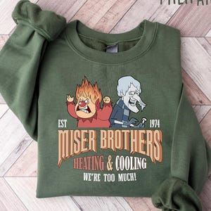 May include: Military green sweatshirt featuring a graphic of two cartoon characters with the text "Miser Brothers Heating & Cooling We're Too Much!" The design includes the year "1974". The sweatshirt is folded on a light wood-grain surface.