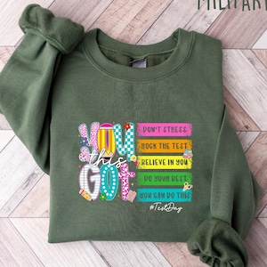 May include: Military green sweatshirt with the words "You Got This" in a colorful, playful font. The design includes stacked books with motivational phrases like "Don't Stress" and "Rock the Test." The hashtag #TestDay is also included.