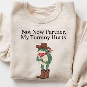 May include: A beige sweatshirt with a green frog wearing a cowboy hat and bandana. The text "Not Now Partner, My Tummy Hurts" is printed on the front.