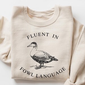 May include: A sand-colored sweatshirt with a black and white illustration of a duck and the text "Fluent in Fowl Language".