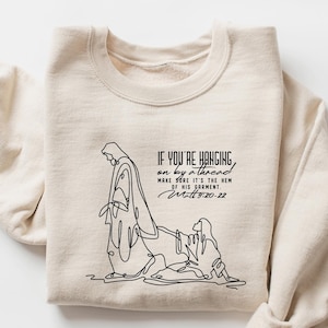 May include: Sand-colored sweatshirt with the text "IF YOU'RE HANGING on by a thread MORE FRE IT'S THE HIM OF HIS GARMENT Matt 20-22" and a line drawing of a religious scene. The word "SAND" is in the upper right corner.