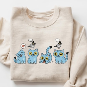 May include: A beige sweatshirt featuring a playful design of four blue cartoon cats with yellow eyes and black stripes. Each cat has a bird perched on its head. One cat has a heart speech bubble, while others have thought bubbles. The word "SAND" is in the upper right corner.