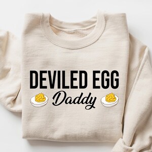May include: Sand-colored sweatshirt with the text "DEVILED EGG Daddy" in black, with two deviled egg illustrations. The sweatshirt is folded, with the sleeves visible. The word "SAND" is in the upper right corner.