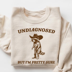 May include: A beige sweatshirt with a brown graphic of a raccoon wearing a cowboy hat. The text "Undiagnosed but I'm pretty sure" is printed on the sweatshirt.