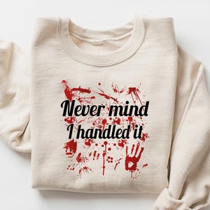 May include: Sand-colored sweatshirt with the text "Never mind I handled it" in black and red. The design includes red splatter and handprints. The sleeves are rolled up.