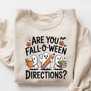 May include: Sand-colored sweatshirt with the text "ARE YOU FALL-O-WEEN DIRECTIONS?" and illustrations of ghosts holding a book, a pencil, and more books. The design includes pumpkins and a bat. The sweatshirt is folded.