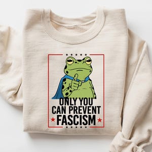 May include: Sand-colored sweatshirt featuring a cartoon frog with a blue cape, giving a thumbs-up. The frog is inside a red-lined box with the text "ONLY YOU CAN PREVENT FASCISM". Stars are above and below the text.
