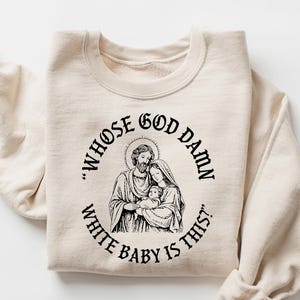 May include: A sand-colored sweatshirt with a black graphic of a religious family and the text "WHOSE GOD DAMN WHITE BABY IS THIS?" The sweatshirt is folded with the sleeves rolled up.