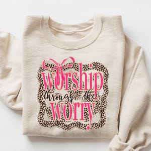 May include: A sand-colored sweatshirt with the text "Worship through the Worry" in pink script and a leopard print border. The sweatshirt has long sleeves and a crew neck.