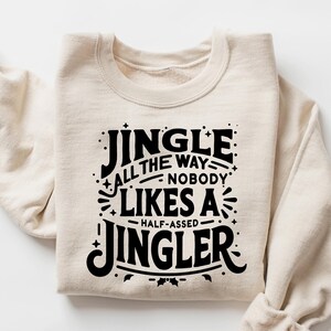 May include: A sand-colored sweatshirt with the text "JINGLE ALL THE WAY NOBODY LIKES A HALF-ASSED JINGLER" in black. The sweatshirt is folded with the sleeves rolled up.