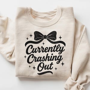 May include: Sand-colored sweatshirt with the text "Currently Crashing Out" in a black script font, accented with a black bow and stars. The sleeves are rolled up, and the overall style is casual and comfortable.