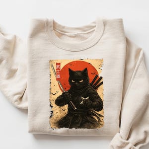 May include: Sand-colored sweatshirt featuring a graphic of a black cat dressed as a ninja, holding a sword and shuriken. The design includes a red sun and Japanese text. The sweatshirt is folded.