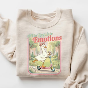 May include: A sand-colored sweatshirt with a graphic of a goose riding a scooter, holding a coffee cup. The graphic is framed with a pink border and includes the text "Off to Regulate Emotions". The sleeves are rolled up.