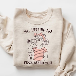 May include: Sand-colored sweatshirt with a graphic of a girl looking through a telescope. The text on the sweatshirt reads "ME, LOOKING FOR WHO THE FUCK ASKED YOU."