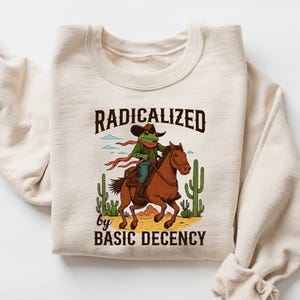 May include: Sand-colored sweatshirt with the text "RADICALIZED by BASIC DECENCY" and a cartoon frog riding a horse. The frog wears a cowboy hat and scarf. Cacti and a desert landscape are in the background.