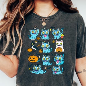 May include: Dark gray t-shirt featuring a Halloween-themed design. The shirt displays a pattern of blue cartoon cats with various Halloween elements like pumpkins, bats, and candy corn. The cats have different expressions and are in various poses.