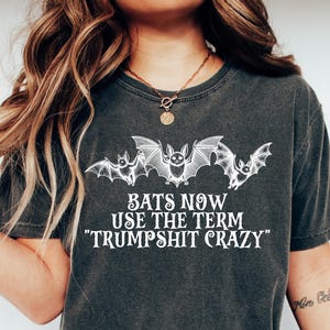 May include: Dark gray t-shirt featuring a graphic of three bats above the text "BATS NOW USE THE TERM 'TRUMPSHIT CRAZY'". The shirt is a casual style, suitable for everyday wear.