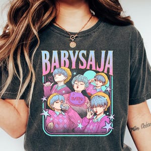 May include: Dark gray t-shirt featuring a colorful graphic with the text "BABYSAJA". The graphic includes anime-style illustrations of characters with blue hair and yellow hats. The shirt is worn by a person wearing blue jeans and white shoes.