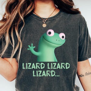 May include: Dark gray t-shirt featuring a green cartoon lizard with pink eyes waving. The text "LIZARD LIZARD LIZARD..." is printed below the lizard in green. The shirt is paired with blue jeans and white sneakers.