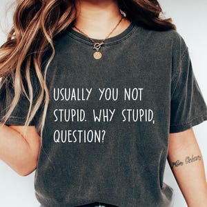 May include: Dark gray t-shirt with the text "USUALLY YOU NOT STUPID. WHY STUPID, QUESTION?" in white. The shirt is paired with blue jeans and white sneakers. A casual, everyday outfit.