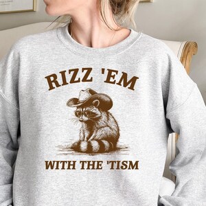K&ouml;nnte beinhalten: Grauer Pullover mit einem braunen Waschb&auml;ren, der einen Cowboyhut und eine Sonnenbrille tr&auml;gt. Der Text "Rizz 'Em With The 'Tism" ist auf dem Pullover aufgedruckt.