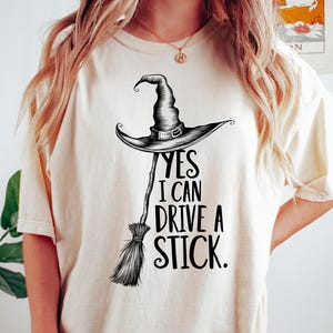 May include: Cream-colored t-shirt with a black graphic of a witch's hat and broom. The text "YES I CAN DRIVE A STICK" is printed below the hat. The shirt is a casual, oversized fit.
