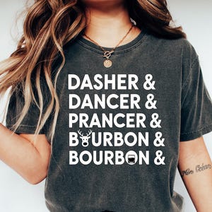 May include: Dark gray t-shirt with white text listing "Dasher & Dancer & Prancer & Bourbon & Bourbon". The shirt is a casual style, suitable for everyday wear. The shirt is paired with blue jeans and white shoes.