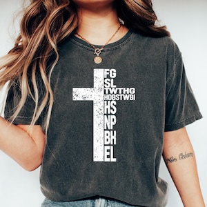 May include: Dark gray t-shirt with a white cross design. The cross is filled with vertical text. The model is wearing blue jeans and white sneakers. The shirt is a casual style.