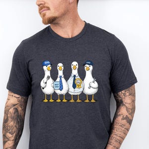 May include: Dark gray t-shirt featuring a whimsical illustration of four cartoon geese. Each goose is dressed in a different outfit, including a cap that says "DAD", ties, and a work uniform. The geese are standing in a row.