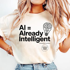 May include: Cream-colored t-shirt with the text "AI = Already Intelligent" in black, along with a brain-shaped lightbulb graphic. The shirt also features the text "powered by curiosity" and a circular logo. The model is wearing light blue jeans.