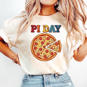 May include: Cream-colored t-shirt with the text "PI DAY" above a pizza graphic. The pizza has a slice cut out, and is topped with pepperoni, mushrooms, and green peppers. The shirt is paired with light blue jeans.