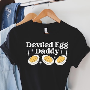 May include: Black t-shirt with the text "Deviled Egg Daddy" and three deviled egg illustrations. The shirt is hanging on a wooden hanger with other clothing items in the background.