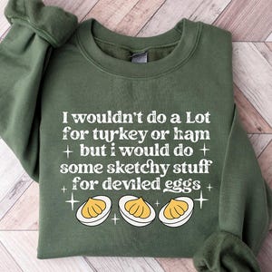 May include: A military green sweatshirt with the text "I wouldn't do a Lot for turkey or ham + but I would do + some sketchy stuff for deviled eggs" and a graphic of three deviled eggs. The sweatshirt is folded on a light wood surface.