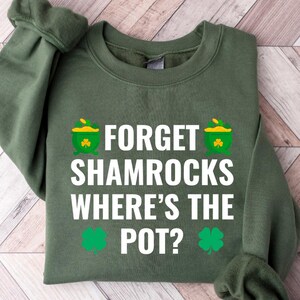 May include: Military green sweatshirt with the text "FORGET SHAMROCKS WHERE'S THE POT?" in white. The design includes two green pots of gold and two green shamrocks. The sweatshirt is folded on a light wood grain surface.