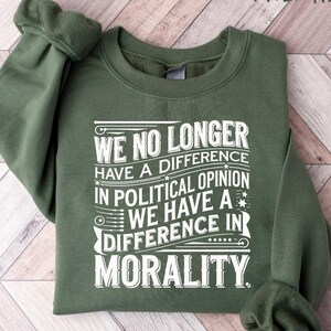 May include: A military green sweatshirt with the text "WE NO LONGER HAVE A DIFFERENCE IN POLITICAL OPINION WE HAVE A DIFFERENCE IN MORALITY" in white. The sweatshirt is folded on a light wood-grain surface.